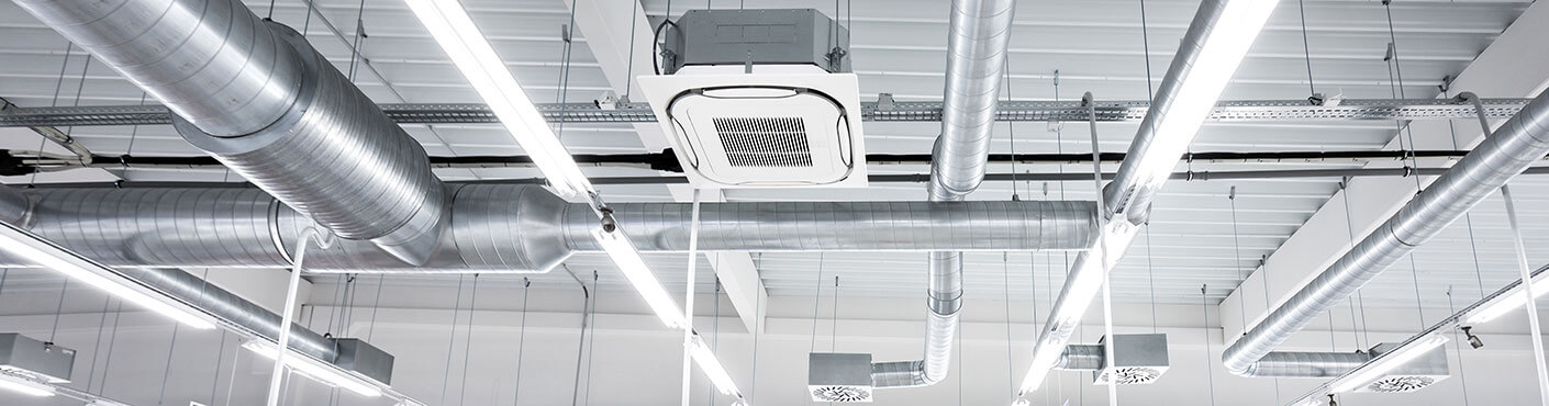 Ceiling mounted cassette type air condition units with other parts of ventilation system (tubes, cables and vents) located inside commercial hall with hanging lights and other construction parts.